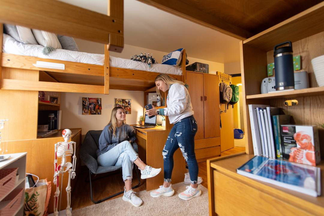 Two young women in a suite style room under a lofted bed. One sits on a chair, and the other stands with a phone.